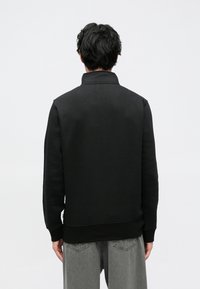 Musta collegepaita, jossa on korkea kaulus, resorit hihansuissa ja helmassa. Valmistettu sileästä materiaalista, istuva malli. Yhdistetty harmaiden farkkujen kanssa.