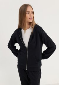 Haut à zip noir en tissu doux, doté d'une poche kangourou, d'une capuche avec cordon de serrage et d'une coupe décontractée, associé à un haut blanc à côtes.