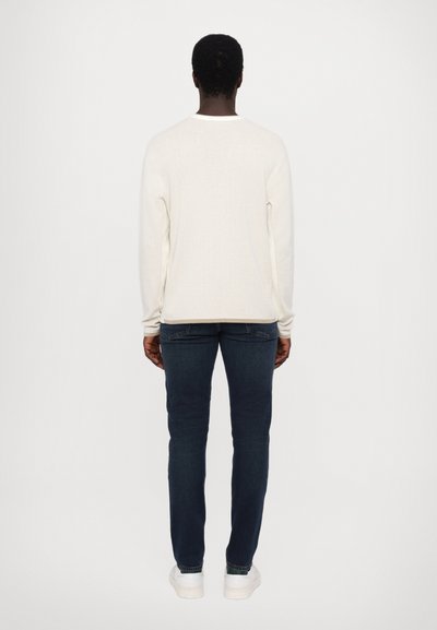 Weißer, gerippter Langarmpullover, Rückansicht, kombiniert mit dunkelblauen Slim-Fit-Jeans und weißen Sneakers vor einem neutralen Hintergrund.