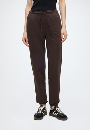 Femme portant un pantalon de jogging marron foncé avec cordon et des baskets noires avec des lacets blancs, debout devant un fond clair uni.
