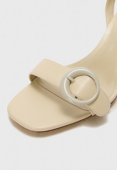 Beige lædersandal med en bred rem og en stor, rund accent, der har en flad sål og en glat tekstur.