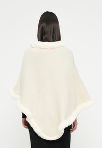 Donna con una capa lavorata a maglia bianco sporco, rifinita con pelliccia bianca lungo il colletto e l'orlo, girata di spalle su uno sfondo semplice.