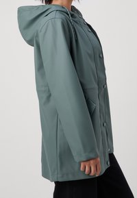 Waterbestendige, teal gekleurde regenjas met capuchon, drukknopen en zijzakken. Heeft een gladde, matte textuur en verstelbare trekkoord.