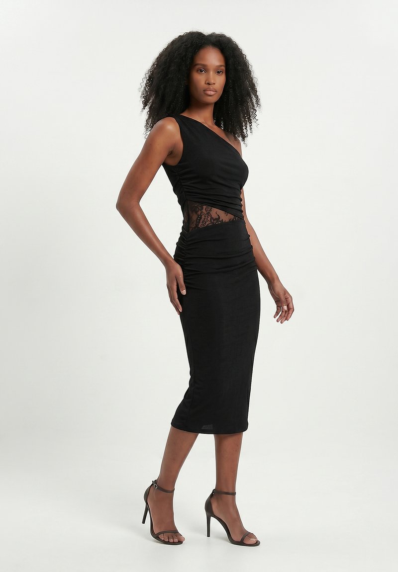 Femme aux cheveux bouclés portant une robe midi noire à une épaule avec une découpe en dentelle à la taille et des sandales noires à talons hauts.