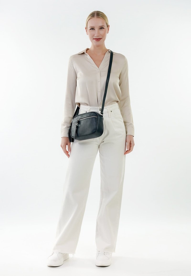 Kvinde iført beige bluse og hvide bukser, står iført hvide sneakers og en sort crossbody-taske mod en ensartet hvid baggrund.