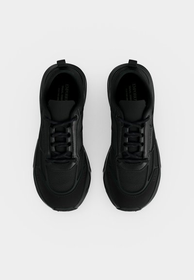 Ζεύγος μαύρα sneakers με κορδόνια και υφή, με λείες επιφάνειες, από πάνω, σε ανοιχτό γκρι φόντο.