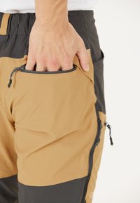 Lichte cargobroek in bruin en zwart met zijritsactiepockets; de stof lijkt glad en duurzaam; hand gestoken in de rechterzak.