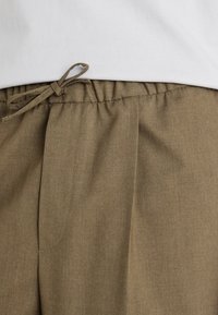 Bruine, gestructureerdebroek met een elastische tailleband en een strik aan de voorkant. Plissé-ontwerpdetails zichtbaar langs de stof.