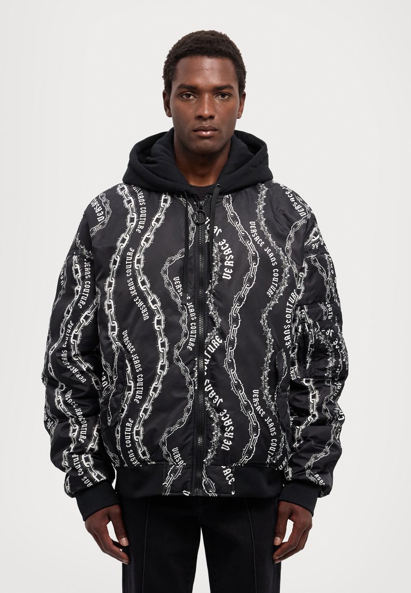 Giacca bomber nera con motivo a catena bianca e testo. Presenta una chiusura con zip, polsini a costine e cappuccio. Realizzata in tessuto lucido.