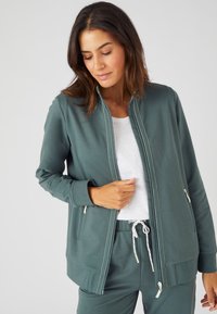 Veste zippée vert foncé en tissu doux, dotée d'un col montant, de poches avant et de détails de couture contrastés en blanc.
