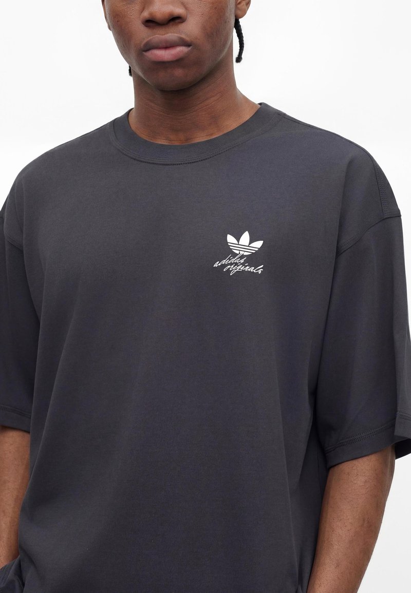 Junge Person trägt ein dunkelgraues Adidas Originals T-Shirt mit weißem Trefoil-Logo und Schriftzug auf der linken Brust vor einfarbigem Hintergrund.