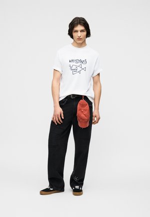 Νέος άνδρας που φοράει λευκό T-shirt με σχέδιο, φαρδιά μαύρη παντελόνια με κόκκινο καπέλο κλιπ στον ζώνη και μαύρα αθλητικά παπούτσια με καουτσούκ σόλες.