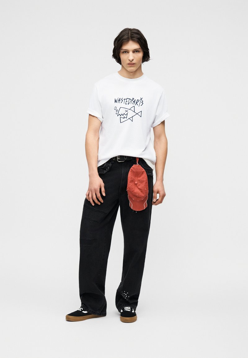 Νέος άνδρας που φοράει λευκό T-shirt με σχέδιο, φαρδιά μαύρη παντελόνια με κόκκινο καπέλο κλιπ στον ζώνη και μαύρα αθλητικά παπούτσια με καουτσούκ σόλες.