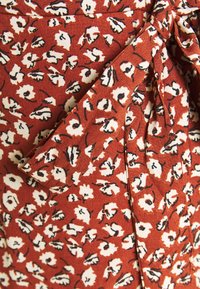 Tissu marron avec un motif floral en blanc et noir, orné d'un détail noué. La texture semble lisse avec une légère brillance.