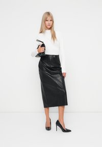 Lange zwarte leren rok met een hoge taille, gecombineerd met een aansluitend wit shirt met lange mouwen. Gekenmerkt door zwarte hoge hakken en een kleine zwarte clutch.