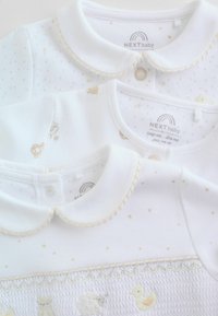 Trois vêtements blancs pour bébé avec des détails de col. L'un présente des animaux brodés ; les autres ont des motifs texturés et à pois avec des accents dorés subtils.