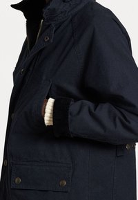 Chaqueta azul marino con cierre frontal de cremallera, botones a presión y bolsillos laterales. Las muñecas de corduroy texturizado y el cuello alzado añaden un toque de detalle.