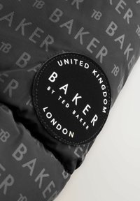 Crni materijal s ponavljajućim uzorkom BAKER logotipa; ima okruglu oznaku branda s bijelim tekstom koja glasi "BAKER BY TED BAKER, LONDON."