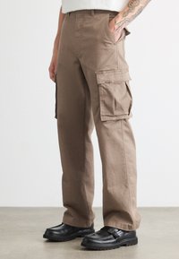Brune cargo bukser lavet af bomuld, med flere sidelommer, et afslappet fit og en glat tekstur. Bæres med sorte slip-on sko.