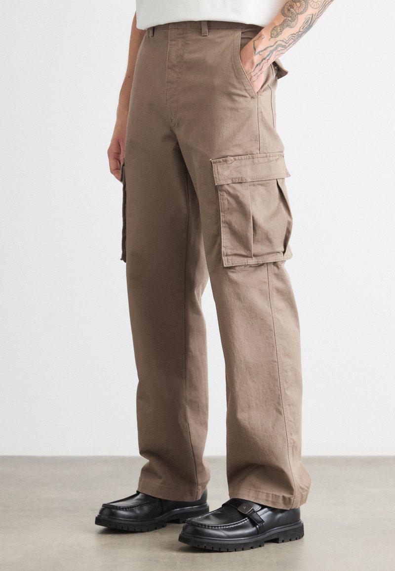 Brune cargo bukser lavet af bomuld, med flere sidelommer, et afslappet fit og en glat tekstur. Bæres med sorte slip-on sko.