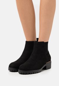 Stivali neri alla caviglia in suede, con suola in gomma spessa, accenti elastici sui lati e una sottile finitura dorata sul tacco.