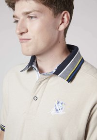 Polo beige avec col rayé bleu marine, doublure intérieure florale, patte à trois boutons et logo brodé sur la poitrine. Matière en mélange de coton doux.