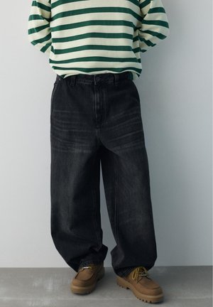 Person iført løstsiddende sorte jeans, brune snørestøvler og en grøn og hvid langærmet trøje med vandrette striber mod en grå væg.