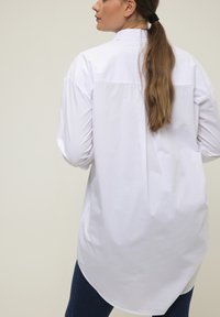 Camisa blanca de manga larga con dobladillo curvado, cuello abotonado y textura de algodón suave, parcialmente metida en unos vaqueros azules.