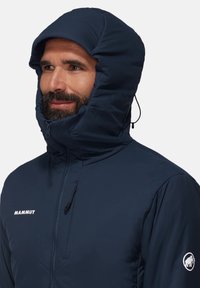 Marinblå isolerad jacka med hög huva, utrustad med dragkedja och snörning, logotyp på bröstet och ärmen, samt en smidig, lätt design.