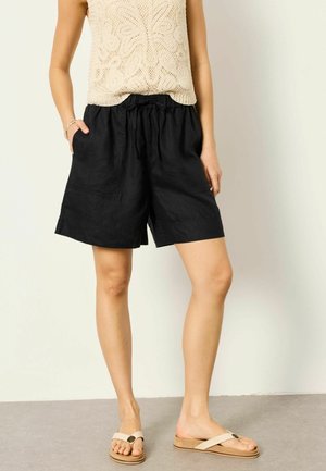 Žena nosiaca čierne šortky sťahovacie na šnúrky, béžový pletený bezrukávový top a béžové sandále s kríženými remienkami, stojaca pri svetlej stene.