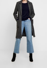Lungo cappotto grigio realizzato in un tessuto testurizzato, abbinato a un maglione blu scuro e a jeans svasati azzurri chiari. Stivaletti neri alla caviglia completano l'outfit.