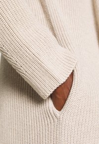 Pull en tricot beige avec texture côtelée, manches longues et une poche latérale, présentant une finition lisse et un design ample.