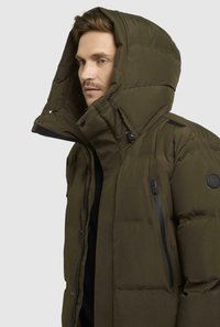 Giacca a vento verde oliva con colletto alto e grande cappuccio. Presenta cerniere visibili, tasche laterali e una toppa con il logo sul braccio.