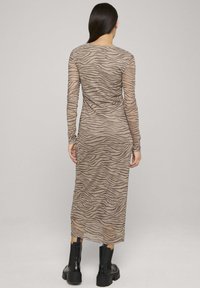 Dolga prosojna obleka z dolgimi rokavi v bež in rjavem zebrinem vzorcu, s prilagojeno silhueto in okroglim izrezom. Oblečena s črnimi robustnimi škornji.