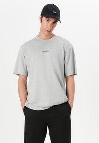 Tricou gri cu mânecă scurtă, cu textură moale și guler cherub; are un mic logo negru „BOSS” imprimat pe piept. Asortat cu pantaloni negri.