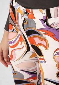 Kleurige broek met een draaiend abstract patroon in oranje, paars, roze en zwart, met een metalen knoop en contrasterende details in de tailleband.