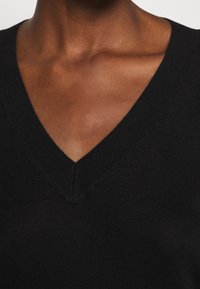 Czarny, dziergany top z dekoltem w serek o prążkowanej fakturze i gładkim wykończeniu. Zbliżenie na linię szyi i obszar barku. Brak widocznych elementów metalowych lub wzorów.