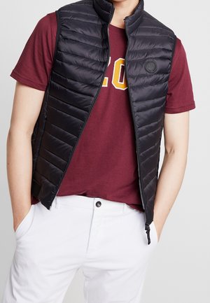 Man draagt een zwart doorgestikt vest over een bordeauxrood T-shirt met gele letters, gecombineerd met een witte broek, handen in de zakken.