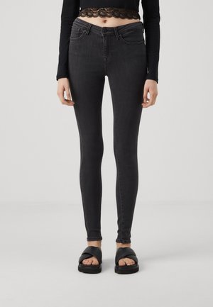 Frau trägt schwarze, hoch taillierte Skinny-Jeans, schwarzes langärmliges Spitzen-Crop-Top und schwarze Plateausandalen und steht vor einem schlichten Hintergrund.