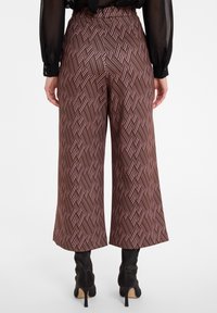 Femme portant un pantalon cropped large à motifs marron et des bottines à talons noires, se tenant avec les mains détendues le long du corps.