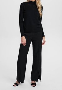 Femme portant un pull noir et un pantalon noir à jambes larges avec des fentes à l'avant, associé à des sandales à talons ouvertes noires, sur un fond uni.