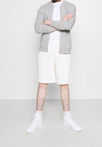 Persona in piedi con le braccia incrociate, indossando una felpa grigia con zip, pantaloni corti bianchi, una maglietta bianca, calze bianche e sneaker alte bianche.