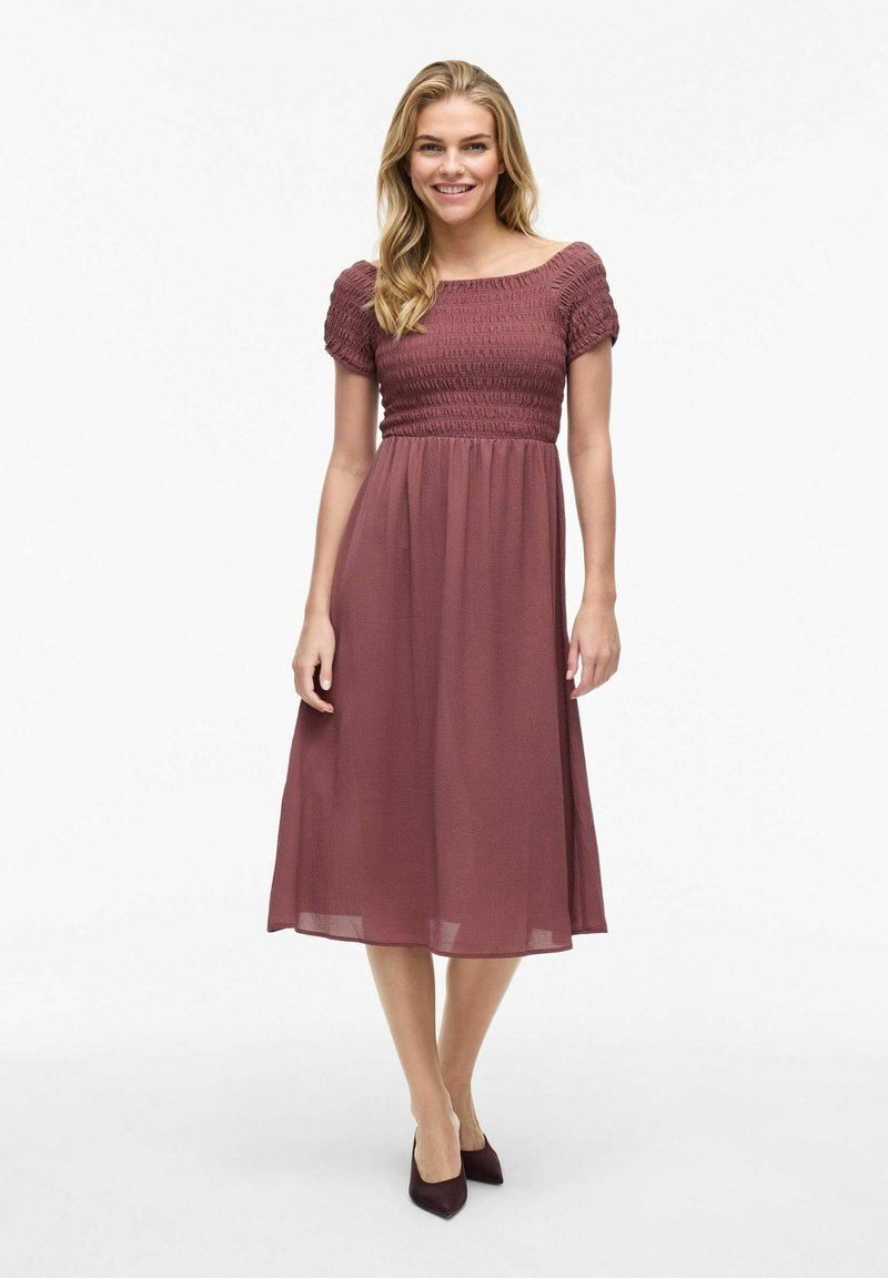 Robe mauve mi-longue avec un corps smocké et des manches bouffantes, fabriquée en tissu léger ; associée à des escarpins noirs à bout pointu.