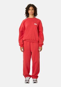 Sarkans, pārāk liels džemperis ar ribotām piedurknēm un apakšmalu, kopā ar atbilstošām brīvām biksēm. Materiāls izskatās mīksts un teksturēts.