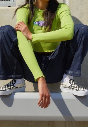 Person trägt ein limettengrünes langärmeliges Crop-Top, dunkelblaue weite Jeans, weiße Socken und schwarz-weiße karierte Slip-On-Schuhe und sitzt mit gespreizten Beinen.