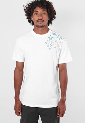 Homme aux cheveux bouclés portant un t-shirt blanc avec des motifs floraux bleus et gris sur l'épaule droite, debout devant un fond uni.