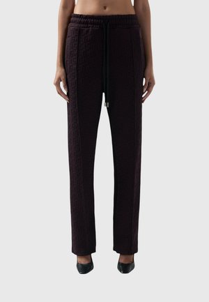 Vrouw die zwarte textuurbroek met hoge taille en trekkoord draagt, gecombineerd met zwarte spitse hakken, staand tegen een effen achtergrond.