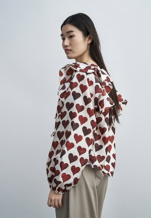 Kobieta z długimi ciemnymi włosami, ubrana w białą bluzkę z czerwonymi wzorami serc i beżowe spodnie, spogląda przez ramię na jednolitym tle.