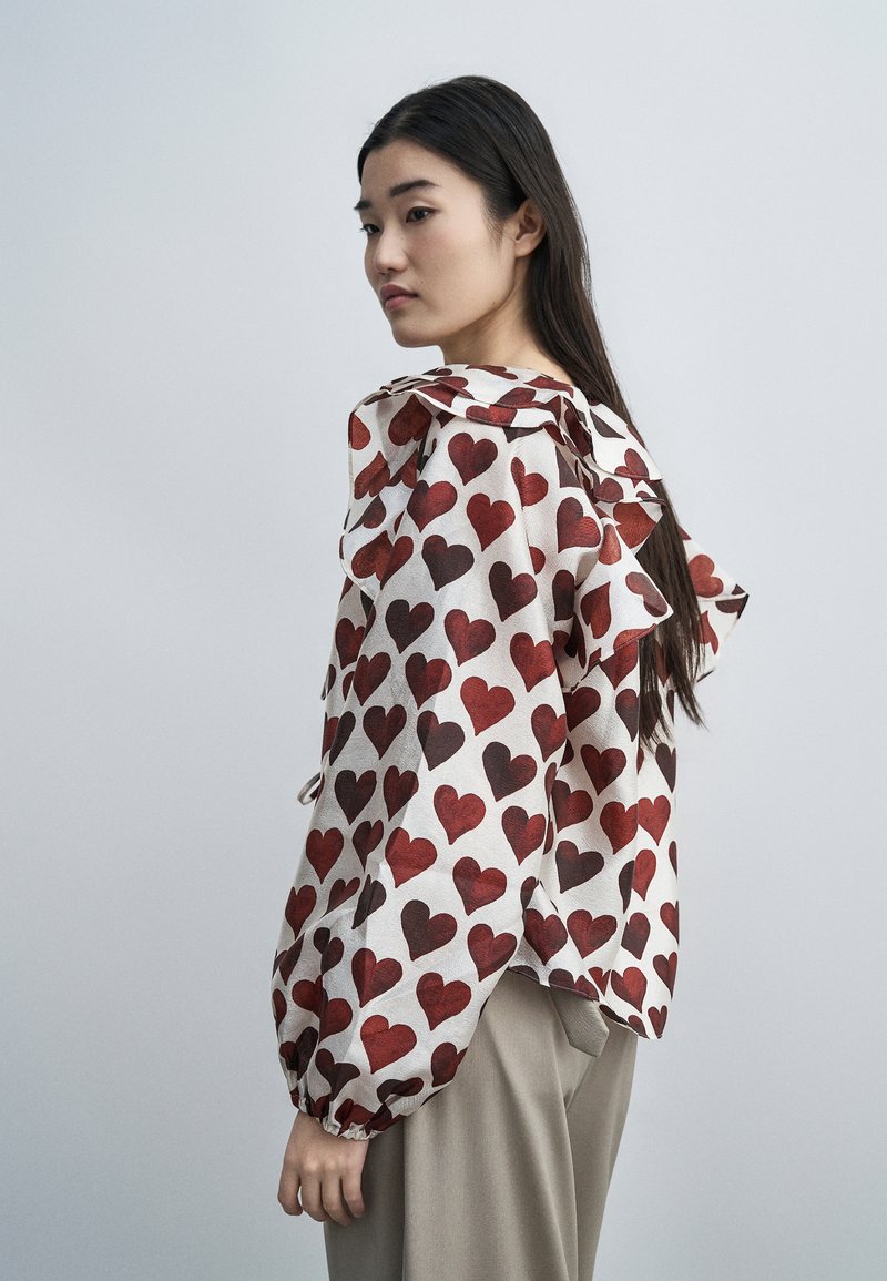 Γυναίκα με μακριά σκούρα μαλλιά, ντυμένη με λευκό πουκάμισο με κόκκινα σχέδια καρδιών και μπεζ παντελόνι, κοιτάζει πάνω από τον ώμο της σε ένα απλό φόντο.