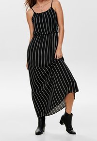 Robe longue noire à rayures verticales blanches, avec fines bretelles, une taille élastique et un ourlet asymétrique. Portée avec des bottines noires.